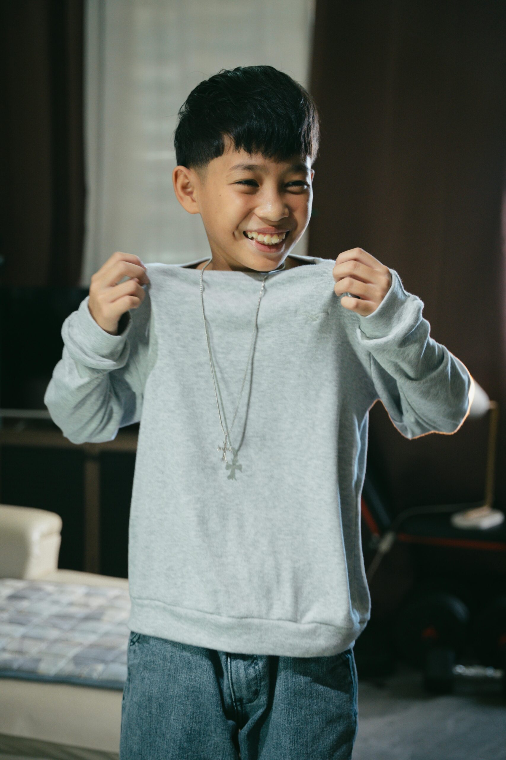 Boy smiling indoors wearing a gray sweater and necklace, conveying joy.