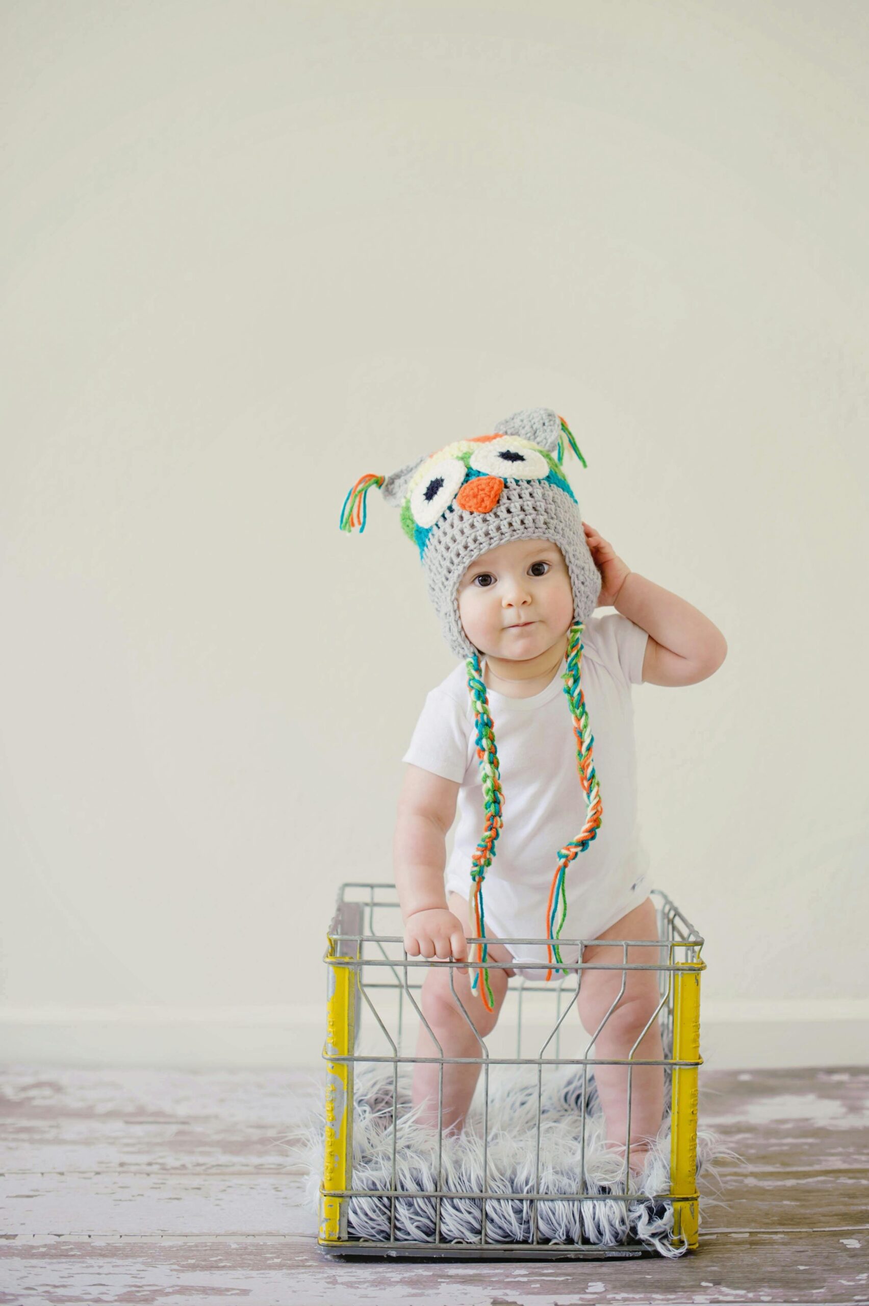 A cute baby wearing an owl hat standing in a basket, smiling and playful.